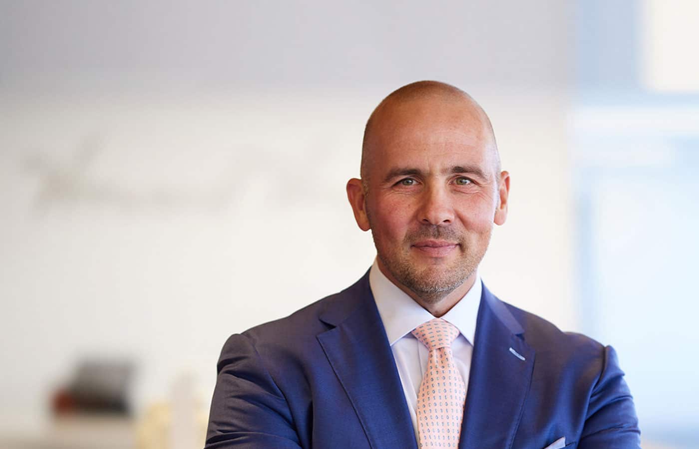 In a vibrant office setting, a bald man confidently stands, wearing a well-fitted suit and tie.